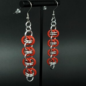 May include: A pair of handmade earrings featuring a chainmail design. The earrings are composed of alternating red and silver-colored metal rings, creating a striking contrast. The earrings hang from silver-colored ear wires.