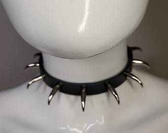 May include: A black leather choker necklace with silver-colored spikes. The necklace is displayed on a white mannequin. The spikes are evenly spaced around the collar.
