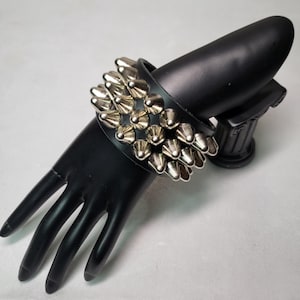 Op de afbeelding: Een zwarte leren armband met zilveren piramidevormige studs. De armband wordt gedragen op een zwarte mannequinhand.