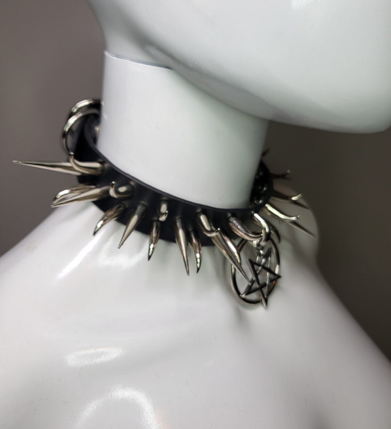 May include: Black leather choker necklace with silver-colored spikes and a pentagram charm. The choker is displayed on a white mannequin, showcasing its punk rock or gothic style. The spikes are evenly spaced around the collar.