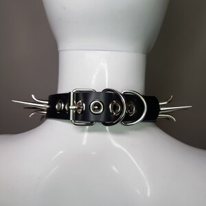 May include: Black leather choker with silver-colored spikes and hardware. The choker features a buckle closure and multiple eyelets for adjustment. The spikes are evenly spaced around the collar, adding a bold, edgy aesthetic.