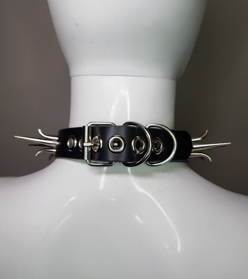 May include: Black leather choker with silver-colored spikes and hardware. The choker features a buckle closure and multiple eyelets for adjustment. The spikes are evenly spaced around the collar, adding a bold, edgy aesthetic.