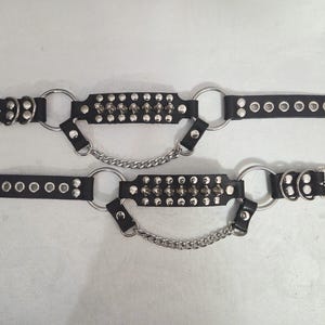 Può includere: Una coppia di polsini neri con borchie metalliche color argento, anelli circolari e una catena centrale di collegamento. Ogni polsino include un cinturino regolabile con occhielli metallici e una fibbia color argento per la chiusura.
