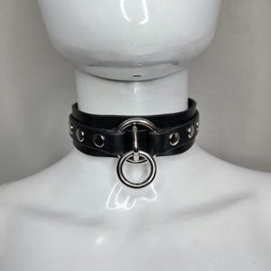 May include: A black choker necklace featuring a wide band with silver-toned metal eyelets and a central O-ring hardware attachment. The design includes a smaller silver ring suspended from the larger central ring, creating a layered metal detail.