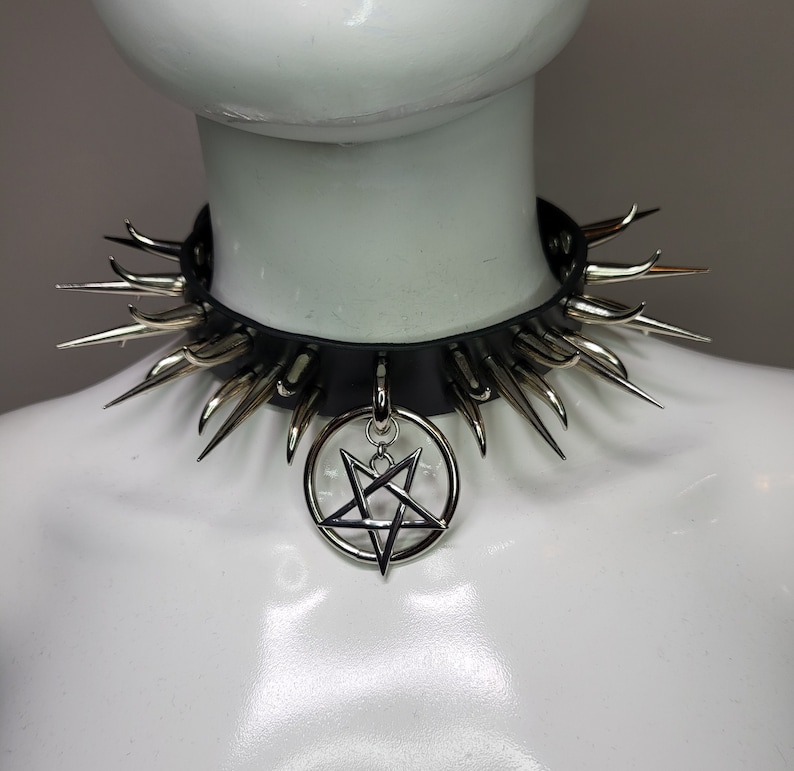 May include: Black leather choker necklace with silver-colored spikes and a pentagram pendant. The choker is displayed on a white mannequin, showcasing the gothic and alternative style of the jewelry.