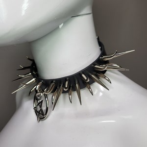 May include: Black leather choker necklace with silver-colored spikes and a dangling pendant. The choker features a series of evenly spaced, pointed metal spikes around the collar. A silver-colored pendant with a star design hangs from the front.