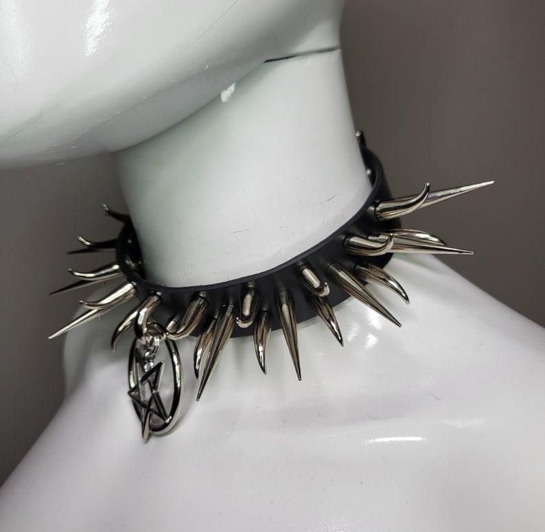 May include: Black leather choker necklace with silver-colored spikes and a dangling pendant. The choker features a series of evenly spaced, pointed metal spikes around the collar. A silver-colored pendant with a star design hangs from the front.