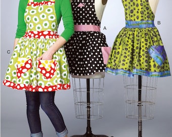 May include: Three aprons displayed on dress forms and a person. Apron C features a green and white egg pattern with red and yellow polka dot pockets. Apron A is black with white polka dots and a pink waistband. Apron B is green with dark circles.