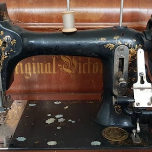 Op de afbeelding: Een vintage zwarte naaimachine met gouden bloemmotieven en de woorden "Original Victor" aan de zijkant. De machine staat op een houten tafel.