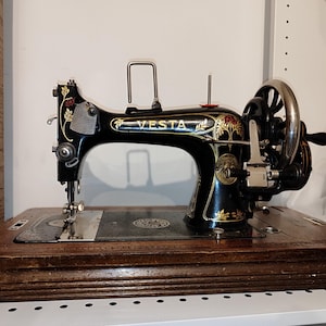 May include: A vintage black sewing machine with gold accents and the word "VESTA" on the front. The machine is on a wooden base with a decorative design.