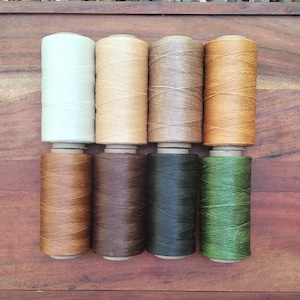 May include: Eight spools of waxed thread in various colours, including cream, tan, brown, and green. The spools are arranged in two rows on a wooden surface, showcasing the different shades available for crafting projects.