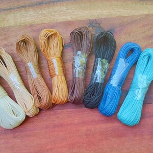 May include: A selection of coloured cords, including cream, tan, brown, black, blue, and turquoise, are displayed on a wooden surface. Each bundle is secured with clear tape. The cords vary in thickness and texture.