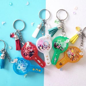May include: Six colorful keychains with different charms and designs. Each keychain features a clear resin charm with various embellishments, including glitter, beads, and small objects. The charms are attached to silver key rings with colorful tassels.