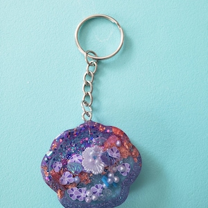 May include: A purple and blue resin keychain shaped like a seashell with glitter, pearls, and other embellishments.