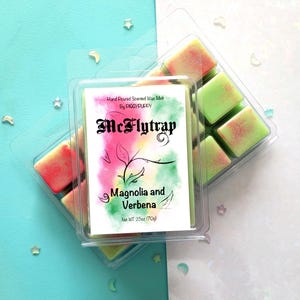 Peut inclure: Deux boîtes en plastique transparent contenant des fondants parfumés. L'une est devant l'autre. La boîte avant porte l'étiquette "McFlytrap Magnolia and Verbena" et "Net WT 2.5oz (70g)". Les fondants sont verts et roses.