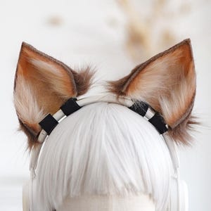 Puede incluir: Un par de orejas de gato de piel sintética marrón y canela unidas a una diadema a rayas blancas y negras. Las orejas están montadas sobre una peluca blanca y unos auriculares blancos. La peluca está sobre una cabeza de maniquí.