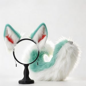 Puede incluir: Un conjunto de accesorios de cosplay con orejas y cola de piel sintética. Las orejas son blancas con bordes turquesa y detalles rojos en el interior, unidas a una diadema negra. La cola es turquesa y blanca.