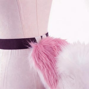 Op de afbeelding: Een roze en witte imitatiebontstaart bevestigd aan een donkerbruine elastische tailleband. De staart is pluizig en heeft een verloop van roze naar wit. De tailleband wordt op een mannequin gedragen.
