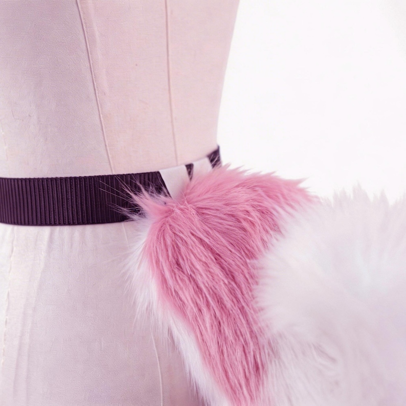 Op de afbeelding: Een roze en witte imitatiebontstaart bevestigd aan een donkerbruine elastische tailleband. De staart is pluizig en heeft een verloop van roze naar wit. De tailleband wordt op een mannequin gedragen.