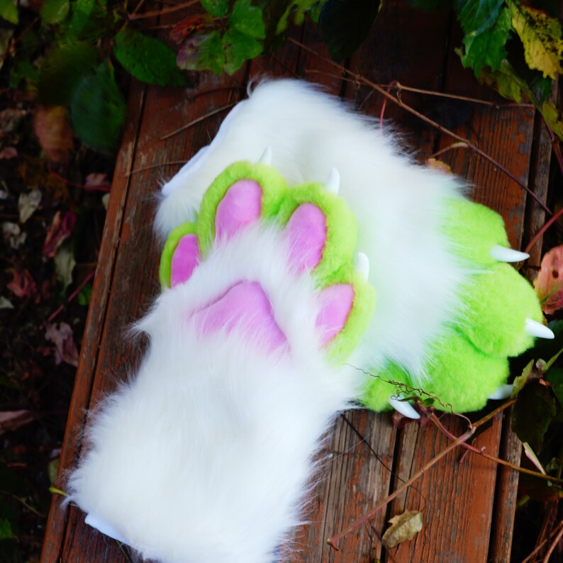 May include: A pair of white and green furry paw hand puppets with pink paw pads. The puppets are lying on a wooden surface.