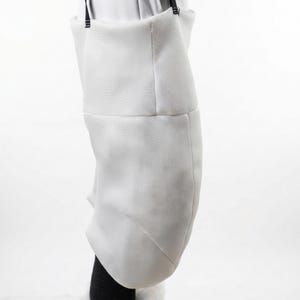 May include: A white fencing jacket, made of a textured fabric, is shown from the waist down. The jacket has a structured shape with a slight curve and is held up by black straps. The lower portion of a black garment is visible.
