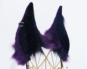 May include: A pair of purple and black faux fur cat ears with silver rings on the left ear. The ears are displayed on a gold metal stand.