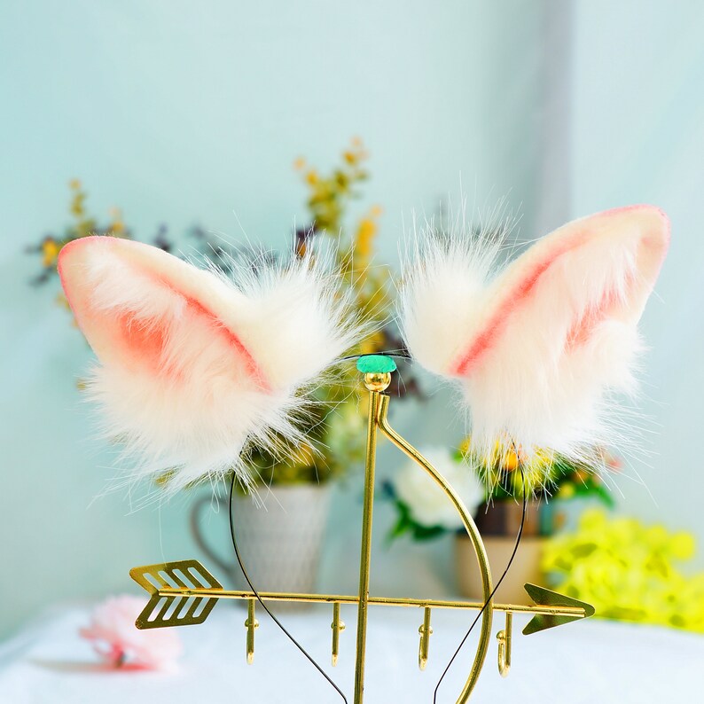 May include: A pair of white faux fur cat ears with pink tips, displayed on a gold stand with an arrow design.