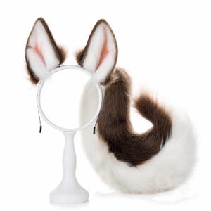 May include: A set of faux fur animal ears attached to a thin white headband, paired with a long, curved faux fur tail. The ears and tail feature a colour pattern of dark brown and white, with soft pink accents visible on the inner ear sections.