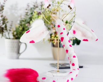 May include: A white and pink spotted tail with a fluffy pink tip, two white and pink spotted ears with pink tips, and a gold headband with pink horns. The tail and ears are displayed on a gold stand.
