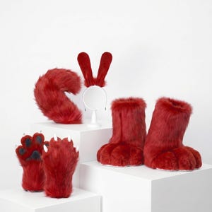 May include: A set of red faux fur costume accessories including a pair of paw-shaped gloves with white claws, a pair of matching furry boot covers, a plush tail, and a headband featuring two tall, upright red ears on a thin metal frame.