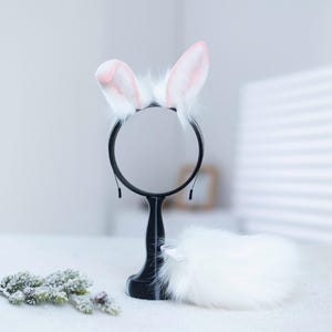 May include: White fluffy bunny ears headband with pink tips, a white fluffy tail, and a black round mirror on a stand.