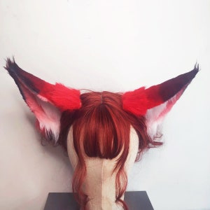 May include: A pair of red and black faux fur fox ears with white tips. The ears are attached to a mannequin head with red hair.