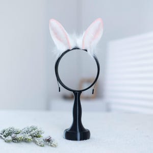 May include: White bunny ears headband with pink tips on a black headband stand. The stand has a round base and a tall, slender stem.