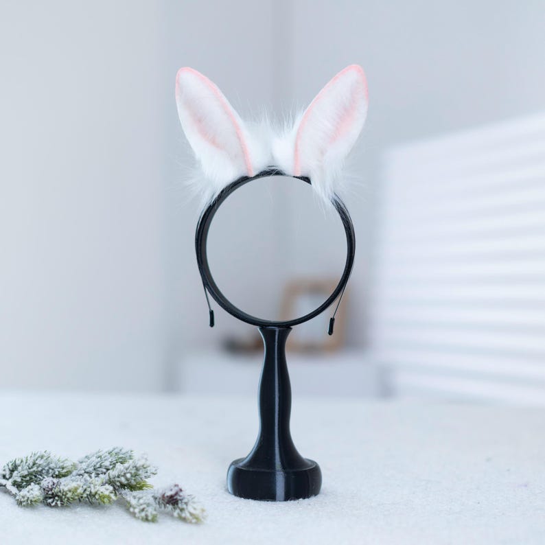 May include: White bunny ears headband with pink tips on a black headband stand. The stand has a round base and a tall, slender stem.