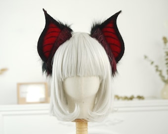 May include: A pair of black and red cat ears with a furry texture. The ears are made of felt and have a red and black striped pattern. The ears are worn on a white wig.