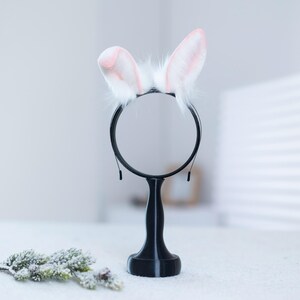 May include: A white bunny ear headband with pink tips on a black stand with a round mirror. The stand is on a white surface.