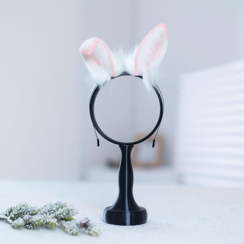 May include: A white bunny ear headband with pink tips on a black stand with a round mirror. The stand is on a white surface.