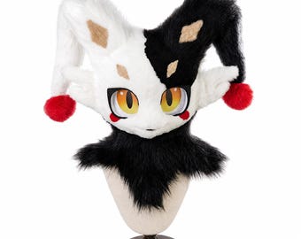 May include: A plush jester-style headpiece featuring a split black and white faux fur design. It includes two long ear-like flaps tipped with red pom-poms, large yellow and orange cat-like eyes, and small tan diamond markings above the eyes.