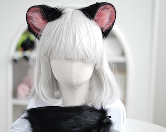 以下が含まれることがあります： 黒とピンクの猫耳カチューシャと、お揃いの黒い尻尾。耳は黒で、内側はピンク色です。尻尾はふわふわで黒色です。白い髪のマネキンの頭に展示されています。