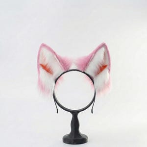 Op de afbeelding: Een zwarte haarband met roze en witte kattenoren van imitatiebont. De oren hebben oranje details. De haarband wordt gepresenteerd op een zwarte standaard.