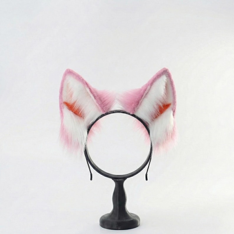 Op de afbeelding: Een zwarte haarband met roze en witte kattenoren van imitatiebont. De oren hebben oranje details. De haarband wordt gepresenteerd op een zwarte standaard.