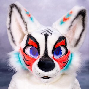 May include: A white fursuit head with black, red, and teal accents. The fox-like face has large blue eyes, a black nose, and detailed markings around the eyes and ears. The fur is fluffy and the overall design is vibrant and expressive.