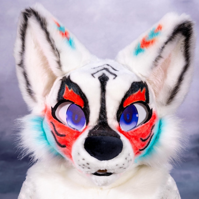 May include: A white fursuit head with black, red, and teal accents. The fox-like face has large blue eyes, a black nose, and detailed markings around the eyes and ears. The fur is fluffy and the overall design is vibrant and expressive.