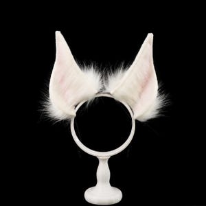 以下が含まれることがあります： 長い白いフェイクファーで覆われた、高く尖った動物の耳が2つ付いたヘッドバンド。耳の内側は淡いピンク色で、縁から毛が外側に広がっており、細いバンドの上に質感のあるふわふわとした外観を作り出しています。