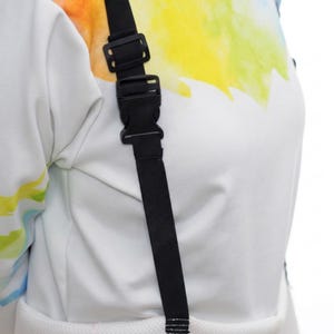 May include: Close-up of a black adjustable strap with a plastic buckle. The strap is attached to a white garment with a colorful, abstract design in shades of yellow, blue, and green.