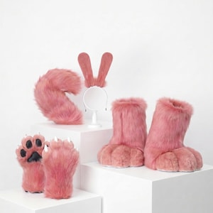 May include: A set of pink faux fur accessories including a pair of paw-shaped gloves with black pads and white claws, a pair of matching furry boots, a fluffy tail, and a headband featuring two long, upright pink rabbit-style ears.