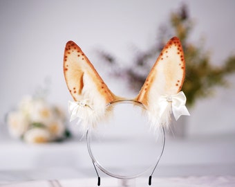 May include: A pair of faux fur ears with brown spots, attached to a headband with white bows and pearl accents. The headband is black and has a white fur trim.
