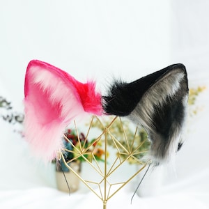 Op de afbeelding: Een paar nep bont kattenoren, een roze en een zwart-grijs, op een gouden metalen standaard.
