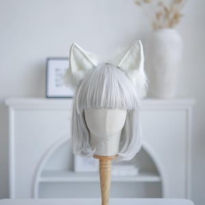 May include: A white, fluffy fox ear headband on a mannequin head with a gray wig. The ears are large and pointed, with a soft, textured appearance. The mannequin is mounted on a wooden stand, set against a white background.
