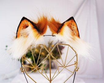 Könnte beinhalten: Ein Paar flauschige Fuchsohren mit einem weißen und orangefarbenen Fellmuster. Die Ohren sind an einem Haarreif befestigt und werden auf einem goldenen herzförmigen Ständer präsentiert.